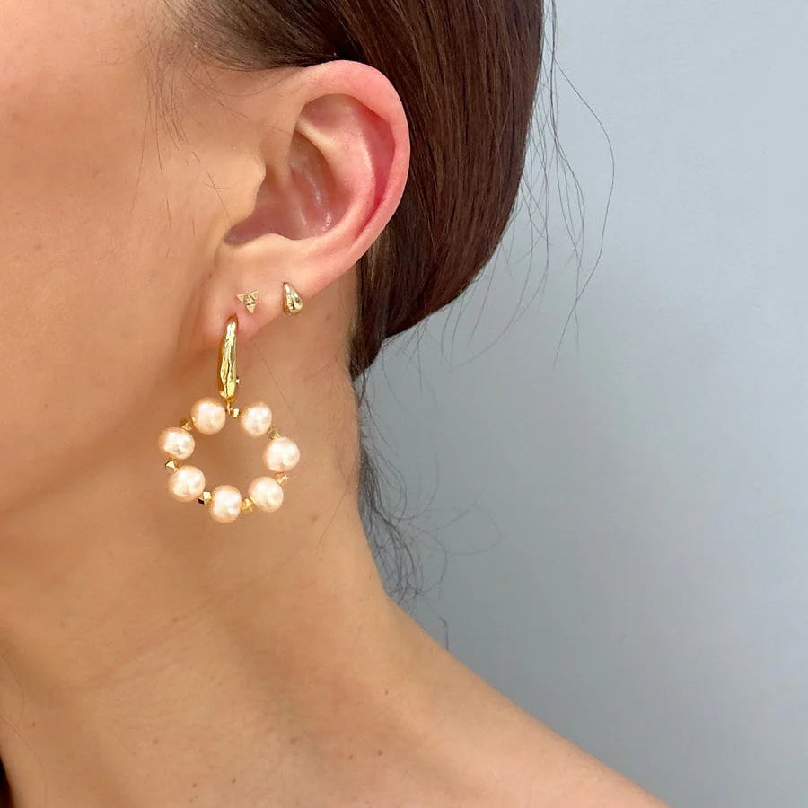 A close-up of a woman's ear displays three Aria V Lunaire Earrings: a small 16k gold stud, a tiny triangular stud, and a large hoop earring with white pearls in a circle, set against a light gray background.