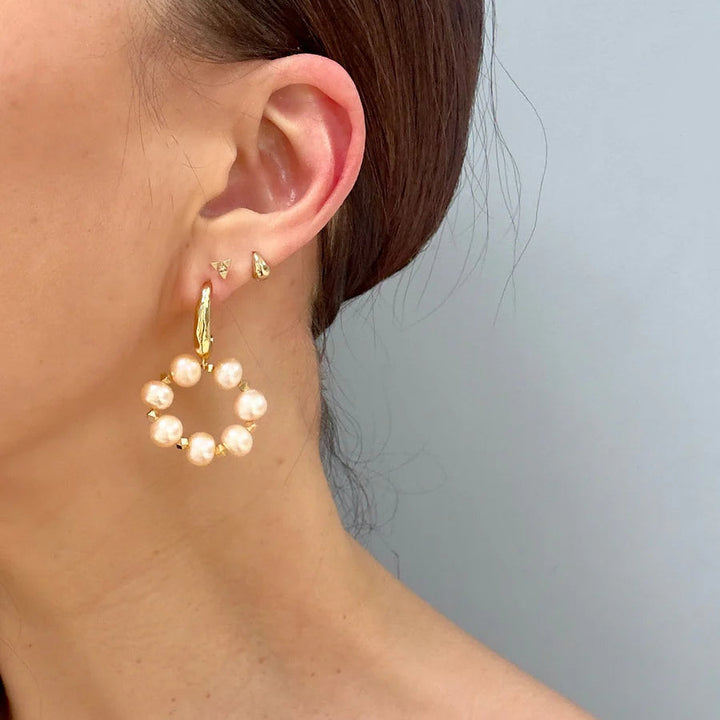 A close-up of a woman's ear displays three Aria V Lunaire Earrings: a small 16k gold stud, a tiny triangular stud, and a large hoop earring with white pearls in a circle, set against a light gray background.
