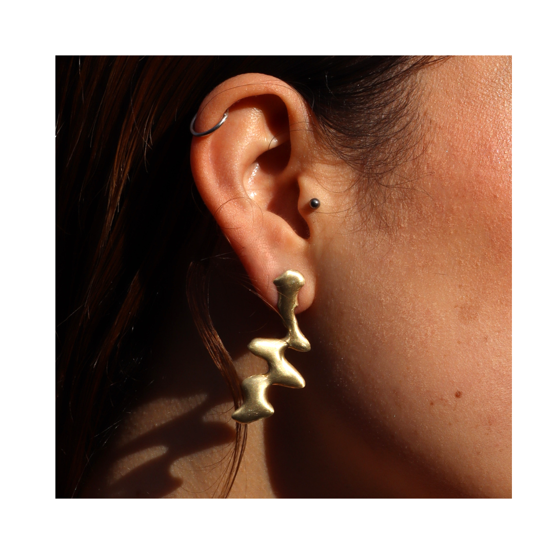 A close-up of a person's ear showcases Blaze Studs by Madeleine Sculptural Jewellery—sculptural earrings crafted from recycled brass, featuring abstract, wavy connected shapes, and illuminated by sunlight.