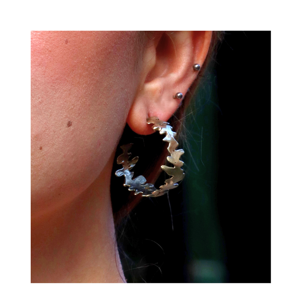 Close-up of a person's ear wearing two small silver studs and a large, jagged-edged Lava Hoop by Madeleine Sculptural Jewellery in the lobe, set against a dark, blurred background.