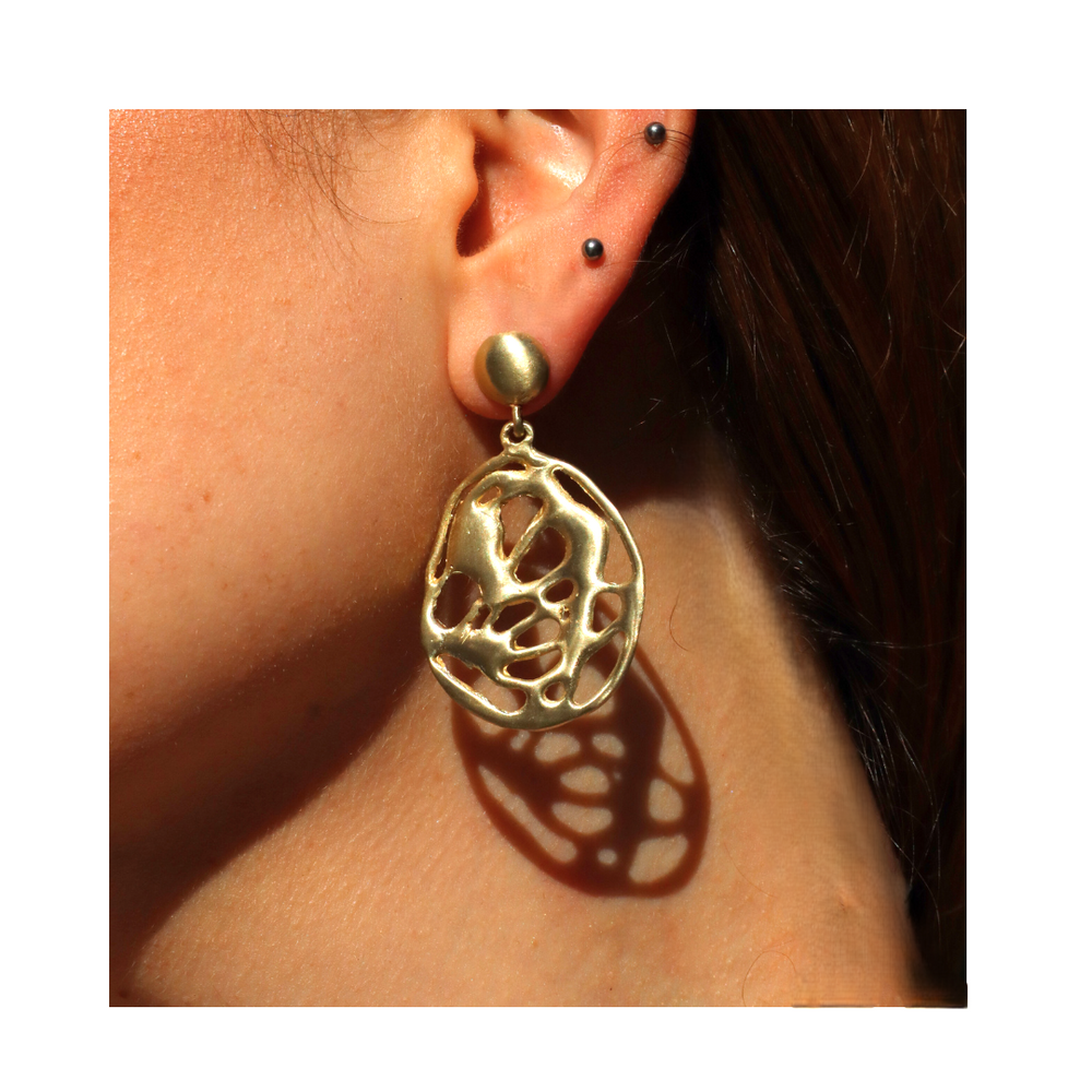 Close-up of an ear with multiple piercings, featuring the Molten Drop Earrings by Madeleine Sculptural Jewellery—large gold, abstract-shaped danglers crafted from recycled brass that cast a decorative shadow on the skin.