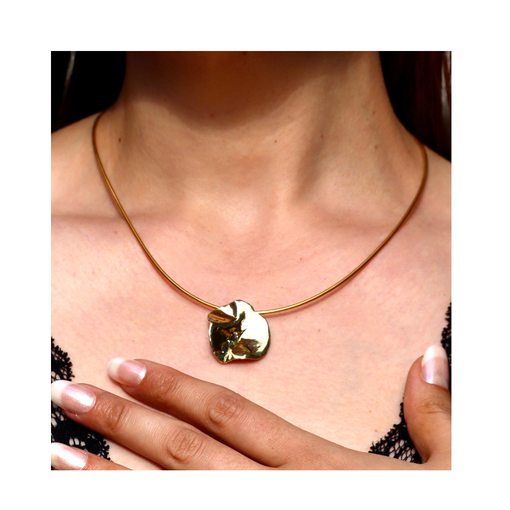 A close-up of a woman wearing the Tide Pendant by Madeleine Sculptural Jewellery— a gold, round, textured necklace— as she touches her chest, highlighting the handmade piece against her black lace top.