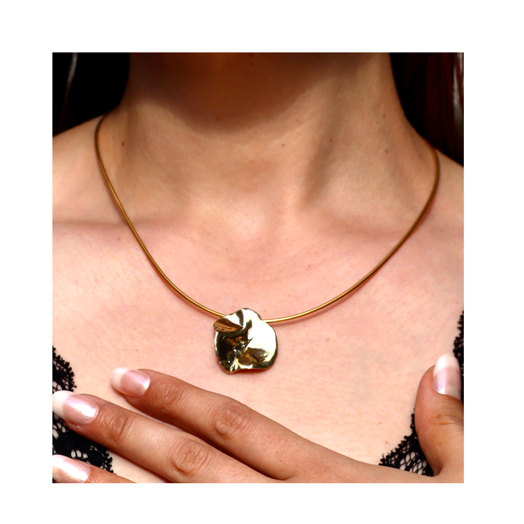 A close-up of a woman wearing the Tide Pendant by Madeleine Sculptural Jewellery— a gold, round, textured necklace— as she touches her chest, highlighting the handmade piece against her black lace top.