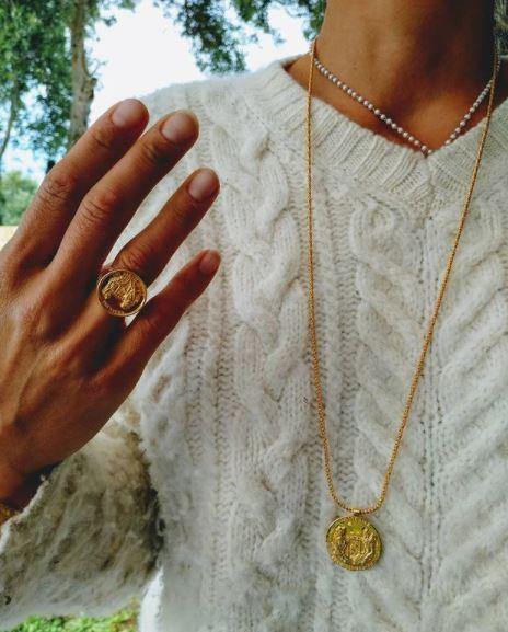 Close-up of a person wearing a cream cable-knit sweater along with the Jennifer Kinnear Grace O'Malley Medallion Small necklace in silver/gold and a matching gold ring. Both the necklace pendant and the ring feature intricate coin designs. The background has some blurry trees and greenery.