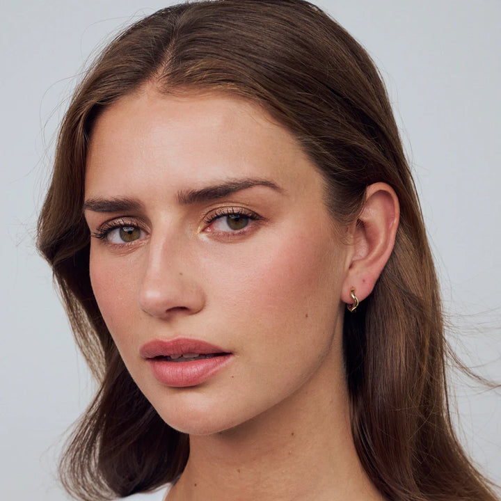 A woman with long brown hair and light makeup gazes at the camera, wearing Sif Jacobs Ellera Waves Pianura Hoop Earrings and a white top, creating a timeless look against a plain light background.