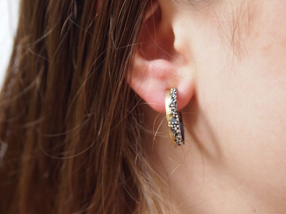 Close-up of a person's ear with straight brown hair, wearing a small gold Etrusc Hoop Earring by MarCucurella, adorned with black and white stones and delicate Etruscan granulation.