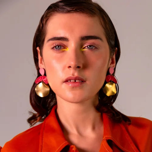 A person with short brown hair wears Shock of Grey's Galaxy Megadrop Earrings in Red and Pink, bold yellow eye makeup, and an orange top, posing against a plain light background.