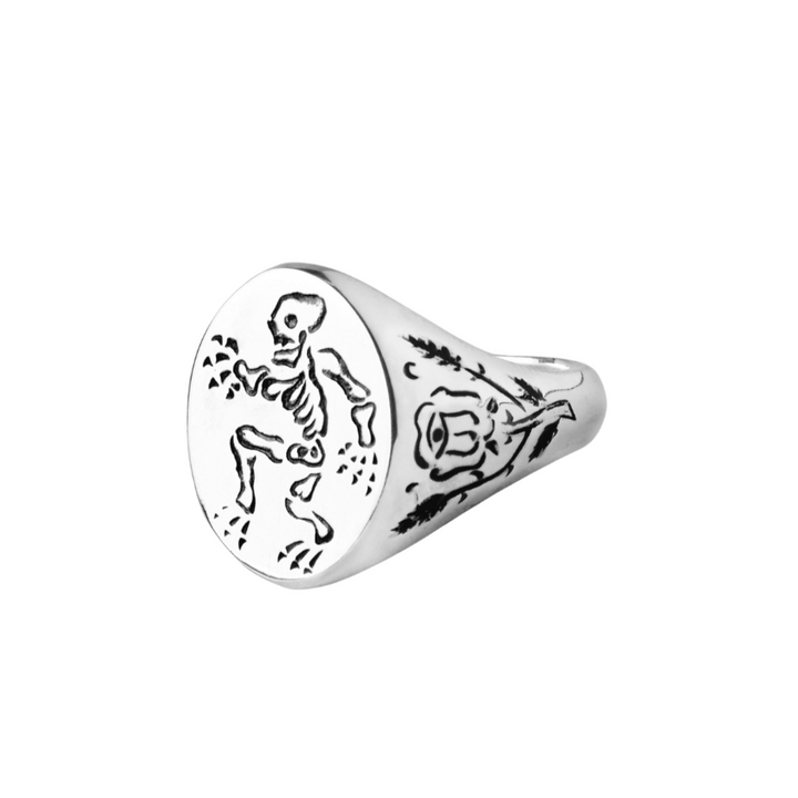 A Cosmic Boulevard Sterling Silver Memento Mori Signet Ring with an engraved skeleton design on the round face, depicting a walking pose. The band is adorned with intricate, hand-engraved patterns and decorative elements, including a stylized rose.