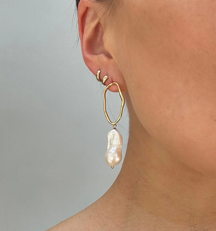 Close-up of a person's ear wearing three small handmade gold Aria V Millie Earrings in various colours on the upper ear and a larger, irregular hoop with a dangling baroque pearl on the lobe, set against a plain background.