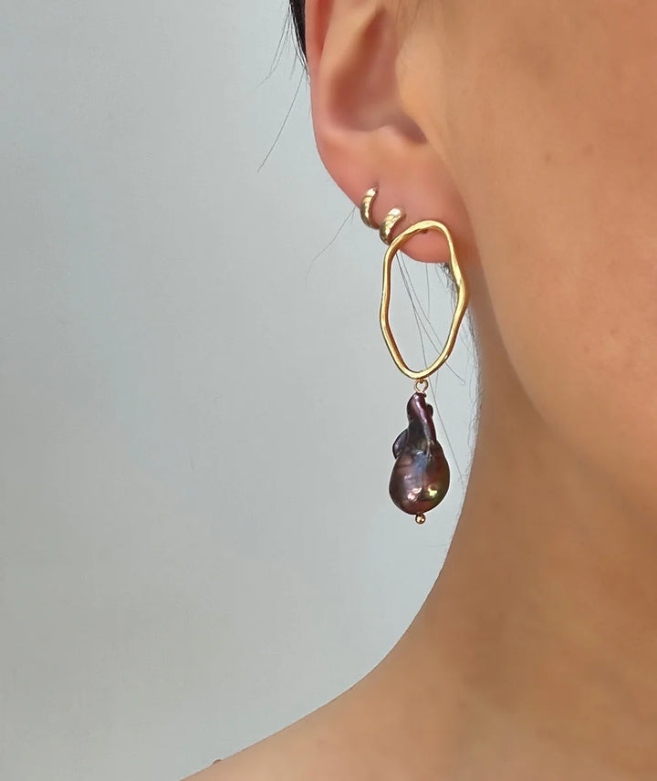 Close-up of a person's ear wearing Aria V's Millie Earrings in various colours, featuring two gold electroplated brass ear cuffs and a large, irregular gold hoop with a dark baroque pearl drop against a plain background.