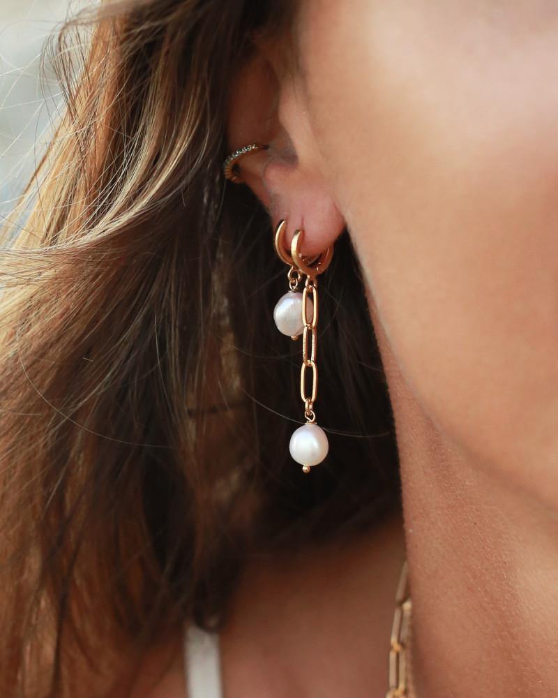 Close-up of a woman's ear adorned with the Nilai Mini Perla Earrings, including a chain-link earring featuring two dangling freshwater pearls and a small hoop earring above it. She has light brown hair cascading down, with loose strands falling near her ear.