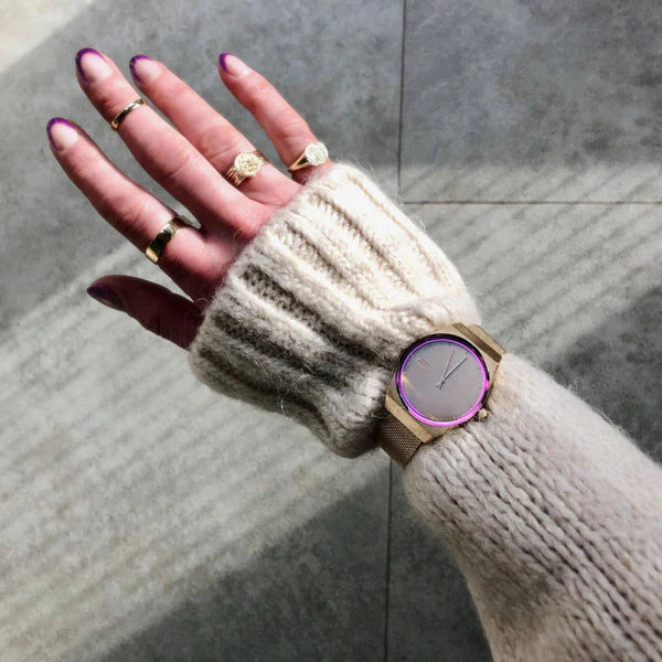 A hand wearing a knitted beige sleeve, adorned with several gold rings, is extended over a tiled floor. The wrist features a Storm Watches Storm Mini Cirero Gold watch with a pink-bordered round face made of stainless steel. The person's nails are painted with a gradient of purple.