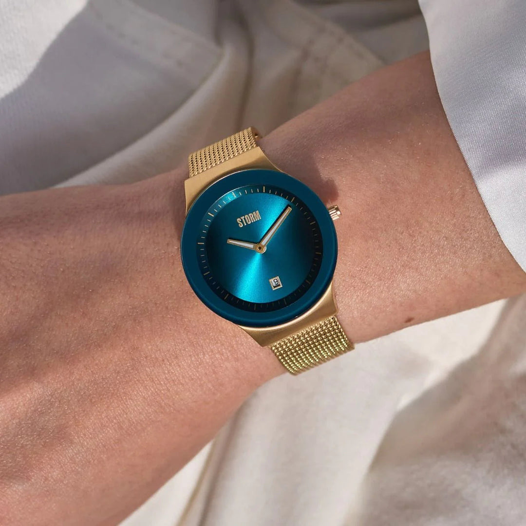 A close-up of a person's wrist wearing the stylish, slim Storm Mini Sotec Gold Teal watch from Storm Watches. It features a turquoise blue dial, gold mesh wristband, and minimalistic design. The edge-to-edge glass showcases thin hands and a small date window. The person is dressed in a light-colored sleeve.