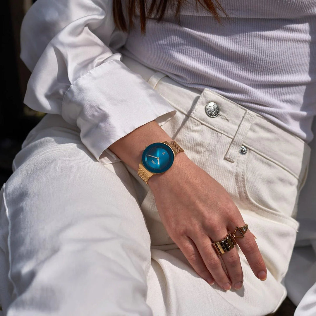 A person wearing a white outfit is seated, showcasing a Storm Watches Storm Mini Sotec Gold Teal wristwatch with edge-to-edge glass and a gold band on their left wrist. Their right hand, adorned with gold rings, rests on their lap. Sunlight casts a warm glow on the scene.