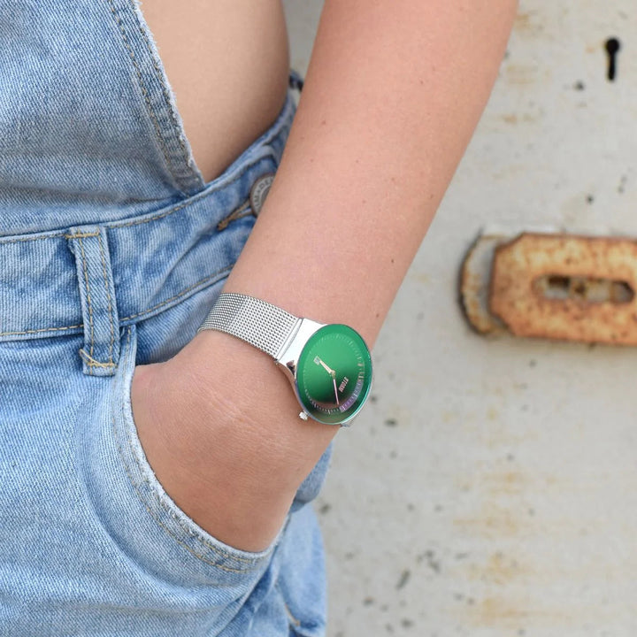 A person wearing blue denim overalls and a green Storm Mini Sotec Lazer Green watch from Storm Watches with a stainless steel mesh strap. The person's hand is resting casually in the front pocket of the overalls while they lean against a rusted, weathered surface.