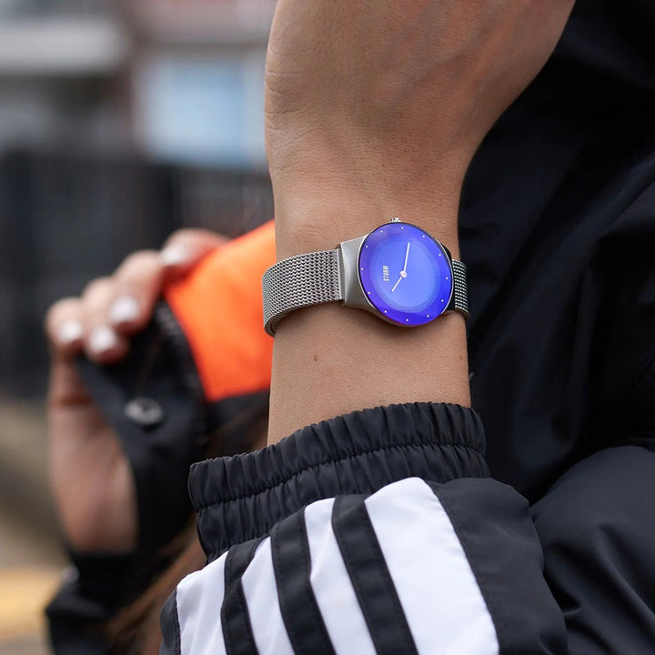 A person wearing a Storm Watches Mini Terelo Lazer Blue watch with a metallic mesh band and minimalist design checks the time. The individual is dressed in a black jacket with white stripes on the sleeves, and an orange accent is visible in the background.