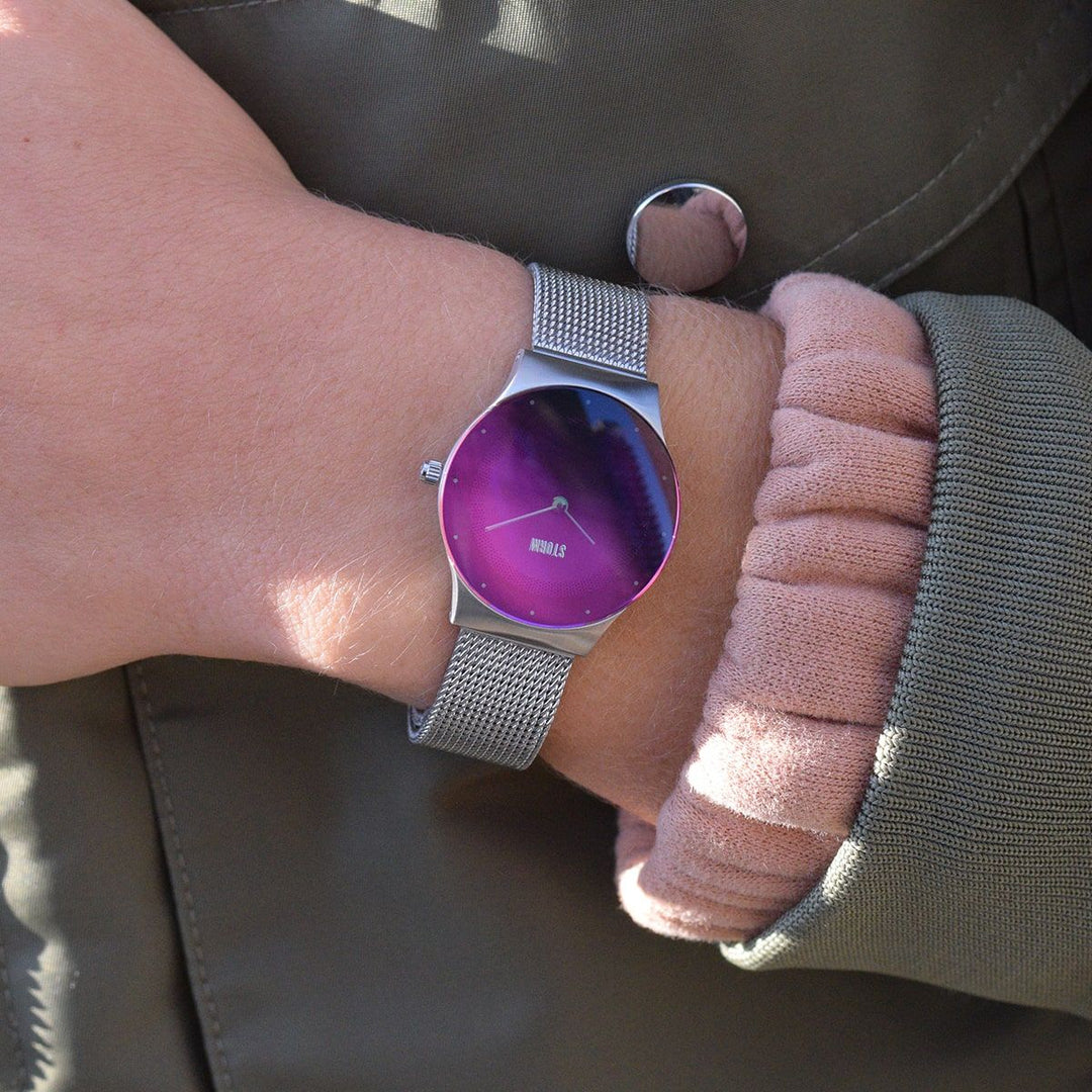 Close-up of a person's wrist showcasing a modern, silver watch with a pink face. The person is wearing a green jacket with a pink cuff peeking out from the sleeve. The Storm Watches Storm Mini Terelo Lazer Purple has a mesh band and minimalist design, resting elegantly on the person's chest.