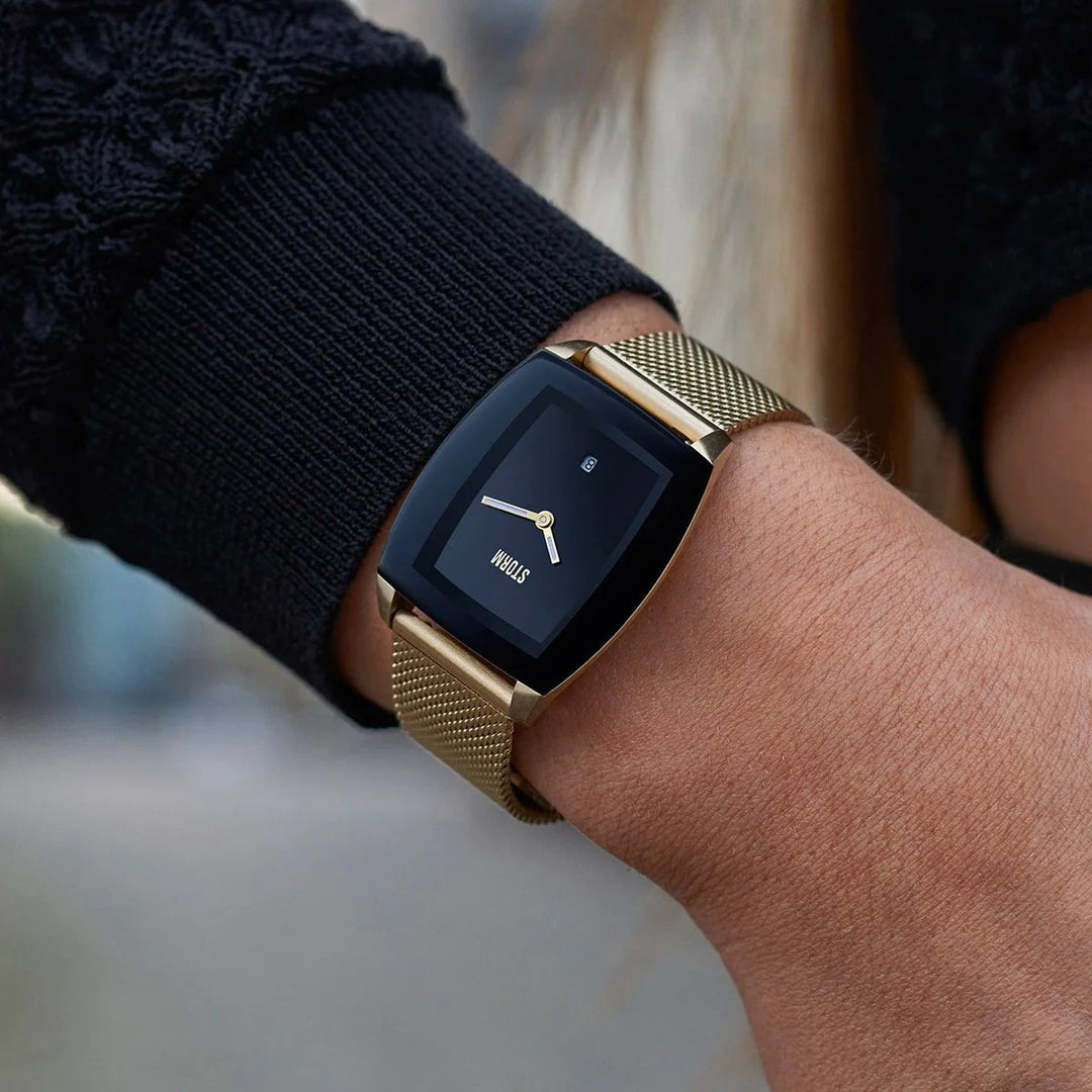 Close-up of a person's wrist wearing a Storm Watches Storm Mini Zaire Gold Black with a black screen displaying a minimalistic clock face and a stylish gold mesh strap. The person is dressed in a dark-colored jacket. The background is blurry.