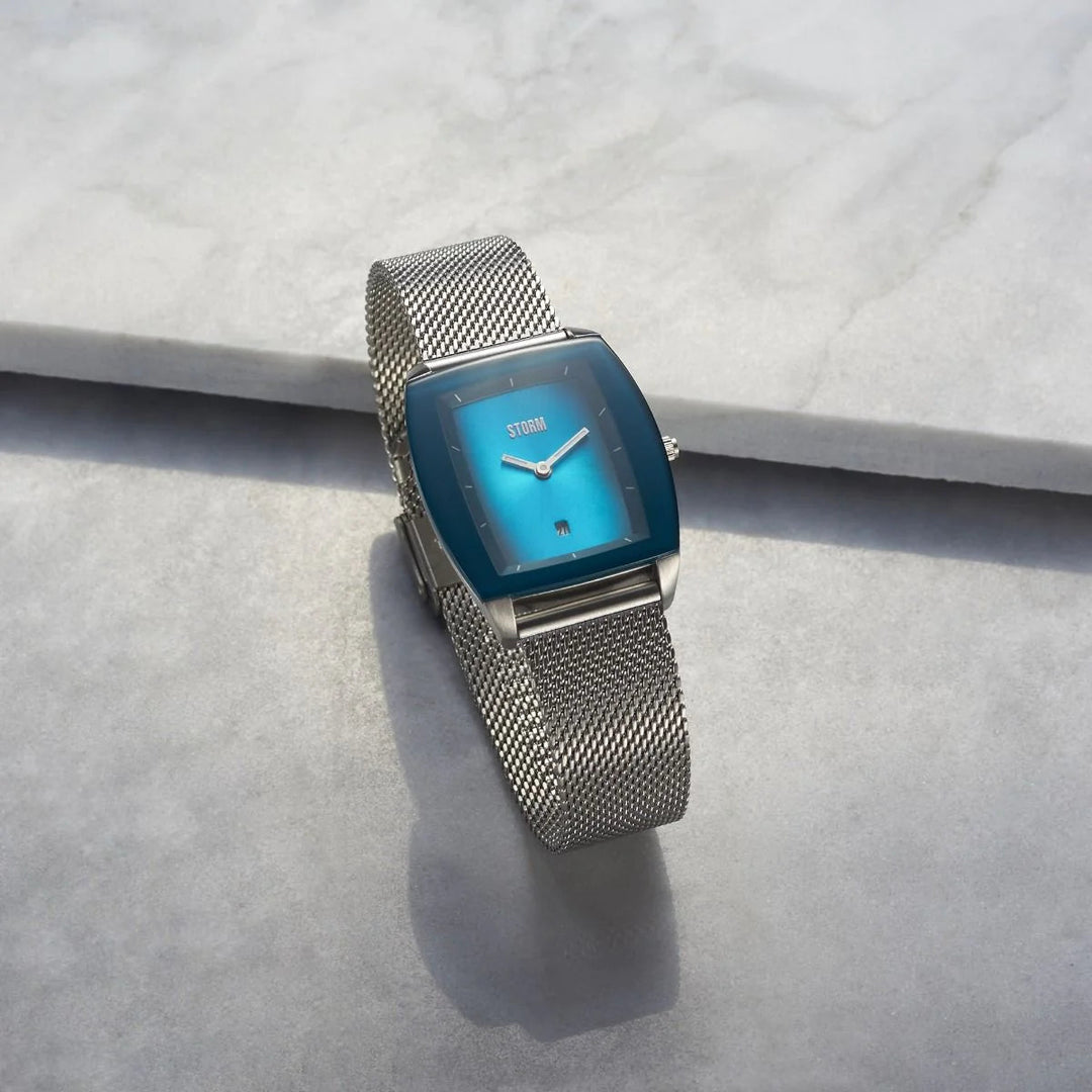 A sleek wristwatch, the Storm Mini Zaire Teal from Storm Watches, features a gold mesh strap and a square blue gradient watch face. Placed on a light grey stone surface, its white hands and minimalist markers contrast against the blue background. The word "STORM" is visible on the watch face.