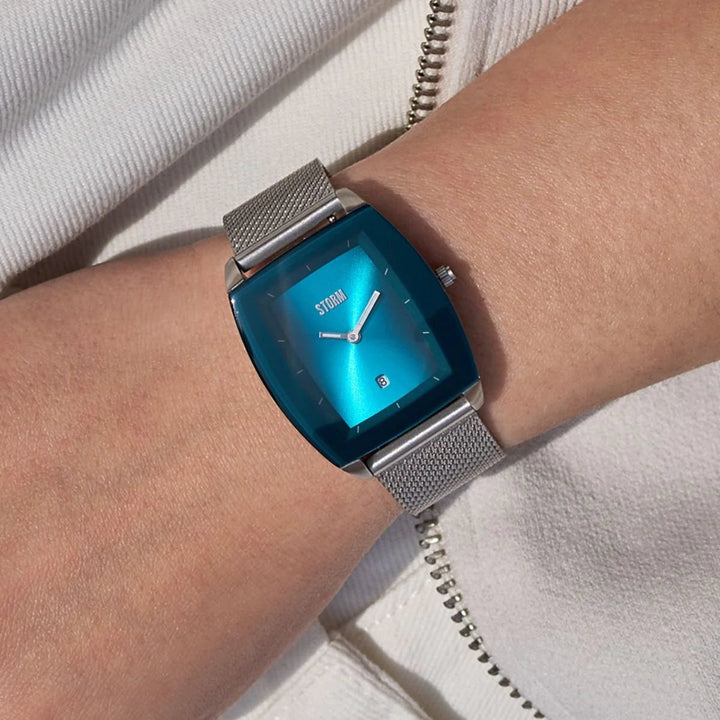 A wrist adorned with a gold mesh strap wristwatch, the Storm Mini Zaire Teal from Storm Watches, featuring a teal-colored square dial displaying the time as 10:10. The hand is resting on a white textured fabric. The watch subtly reflects light, highlighting its sleek design crafted from sustainable materials.