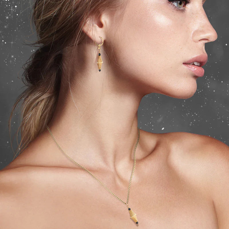A woman with fair skin and blonde hair models the Scribble & Stone Nightsky Cylinder Earrings—14ct gold fill with geometric pendants—paired with a gold necklace, shown in profile against a dark, textured background.