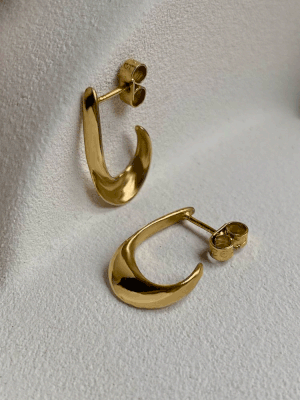 The A Moment Small Earrings by Inner island are small, curved gold minimal earrings with a smooth, shiny finish, displayed against a soft, light-colored textured background.