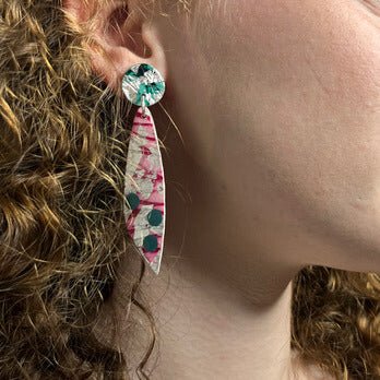A person with curly brown hair wears Rothlú’s Moondrifter Sgraffito Textile Earrings in Silver/Pink/Jade, featuring a round green top and pointed, marbled pink and green dangle. The image focuses on the ear and jawline.