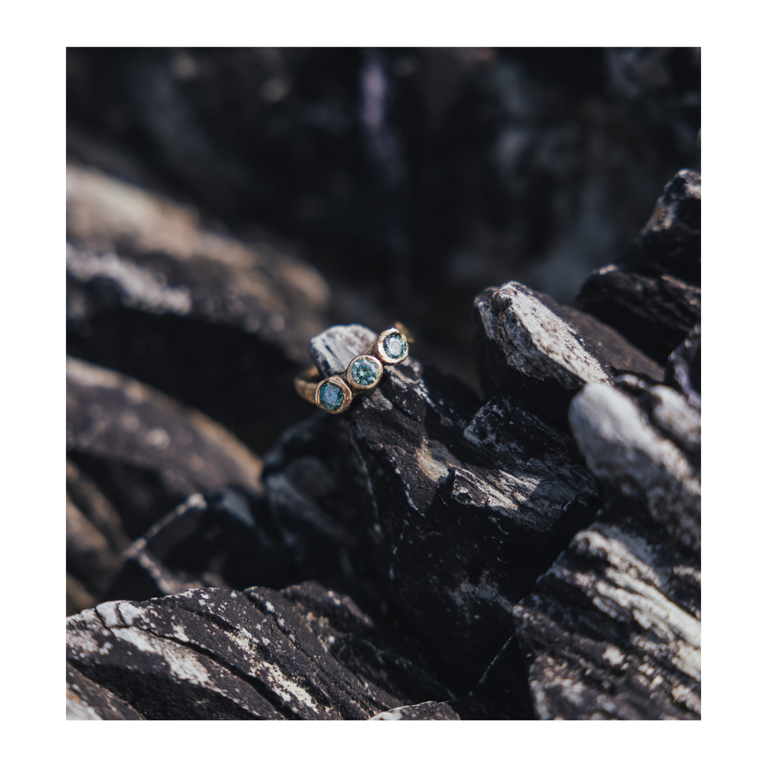 A gold ring with three small round green gemstones, resting on dark textured rocks.