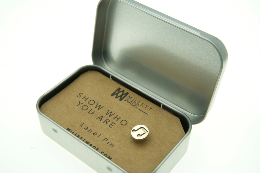 A small, hand-finished brass Music Note Lapel Pin with a musical note symbol is displayed inside an open tin box. The card inside the box reads "Millett Wade - Show Who You Are - Music Note Lapel Pin" with a logo at the top. The box is open, revealing the custom pin and the card.