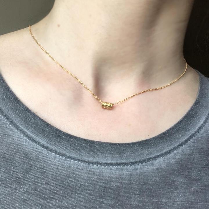 Close-up of a person wearing a simple gold Tiny Karma Wheel Necklace Silver by Andrea Mears Jewellery with a small cylindrical pendant, reminiscent of spinning prayer wheels. The person is dressed in a gray round-neck top. The neutral background perfectly highlights the necklace.