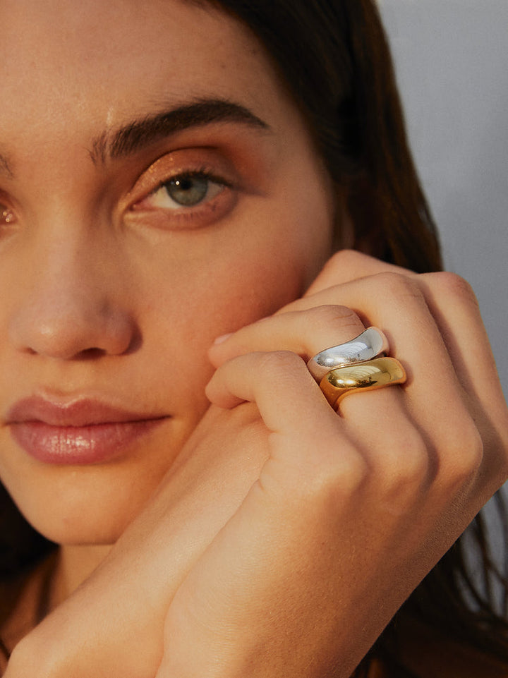 Close-up of a person with light skin and brown hair displaying three stackable Rocco Rings by Shyla on their fingers. The rings are of different colors: silver, white, and gold. The individual has a serene expression, with the focus on their face and the prominent chunky rings.