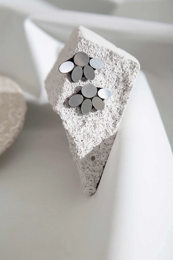 A pair of Organic Reflections Earring Small Silver by Iskin Sisters rests on a piece of textured, white stone, positioned against a soft, neutral background. The design is modern and minimalist, with each earring consisting of overlapping circular petals creating a delicate flower-like form.