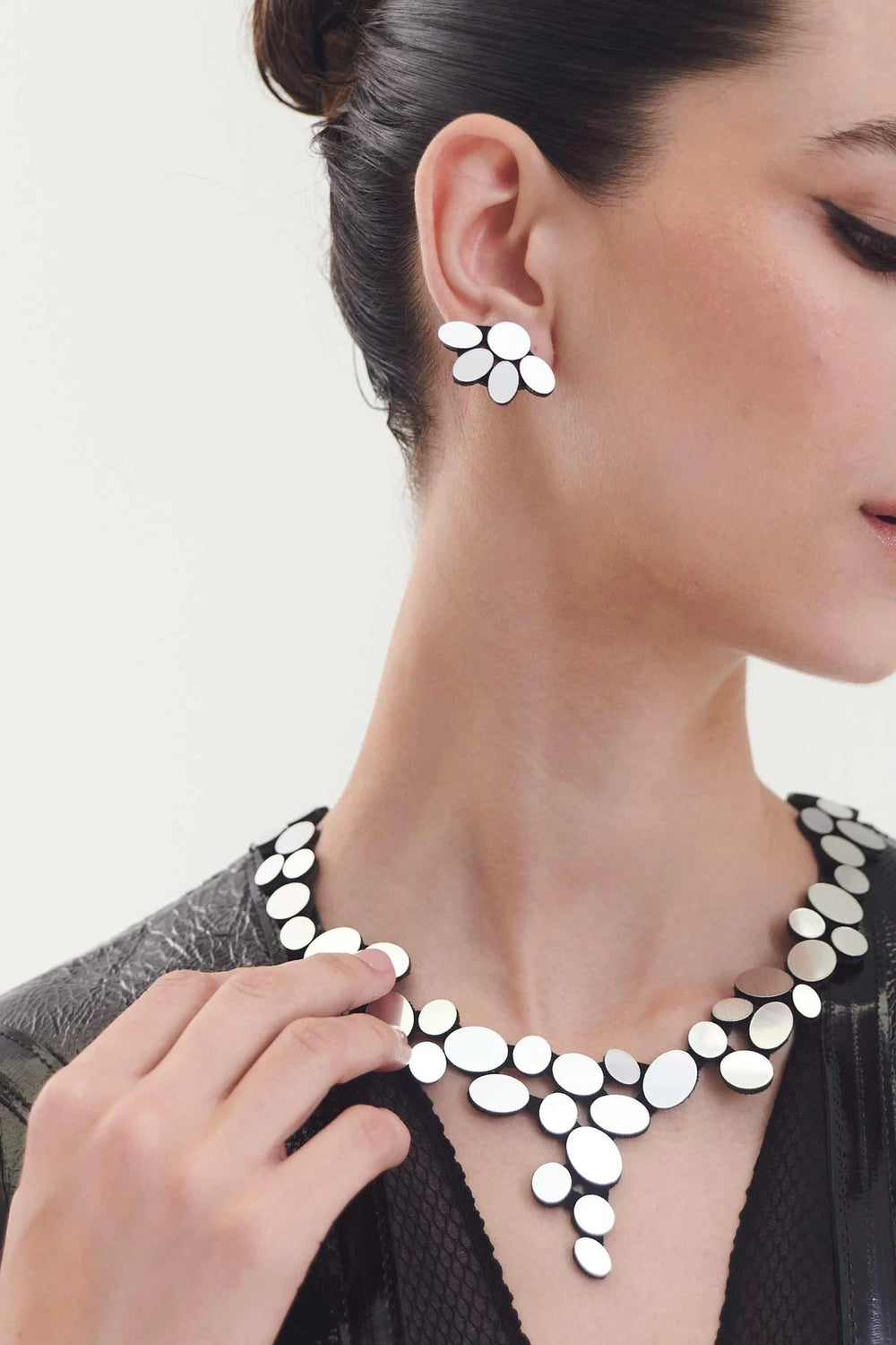 Woman wearing the Organic Reflections Earring Small Silver by Iskin Sisters, along with a necklace made of white oval-shaped pieces. She has short, dark hair tied back and is dressed in a black lace-patterned top. She is touching the necklace with her left hand.