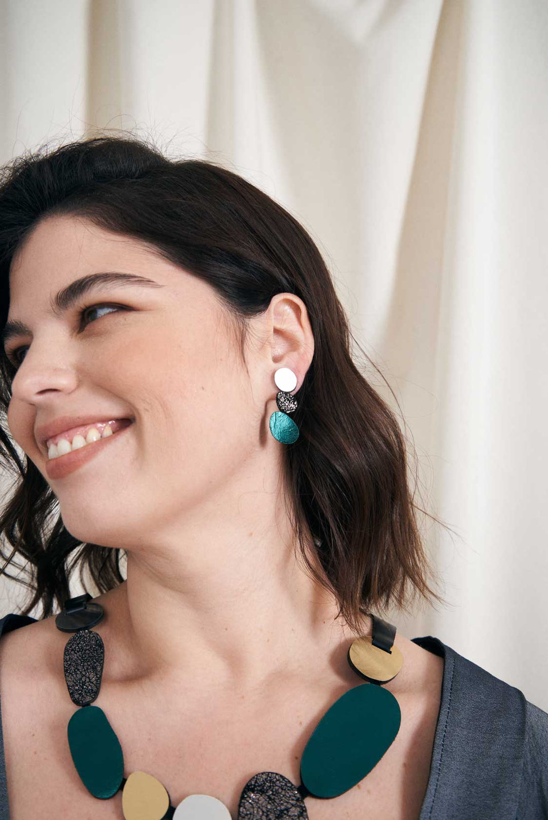 A woman with shoulder-length dark hair is smiling while looking to the side. She is wearing the large, colorful handcrafted Organic Reflections Earrings by Iskin Sisters and a chunky necklace with green, gray, and gold accents. Dressed in a gray top, she stands against a light-colored curtain backdrop.