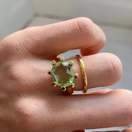 A hand wearing two gold rings: a simple band and Shyla’s Oval Claw Ring in green, featuring a large oval gemstone set in gold.