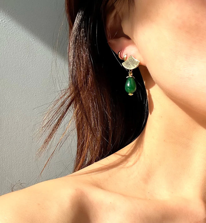 Close-up of a person wearing Pagoda Earrings with a gold and green design by Aria V. The earrings feature a gold fan-like piece at the top and a green jade gem hanging below. The person's ear and part of their neck and shoulder are visible. Light illuminates the skin and earrings.