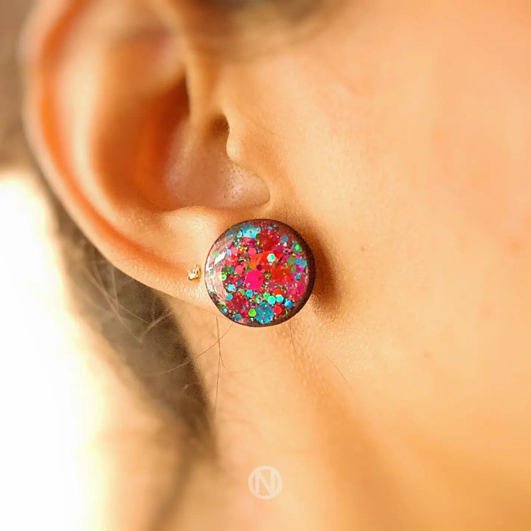 Close-up image of a person's ear wearing Naoi's Party Stud Earrings with a round, colorful design. The earring features multicolour glitter against a dark background. The person has a second small stud earring in gold just above the colorful one, both set on stainless steel earring posts for durability and style.