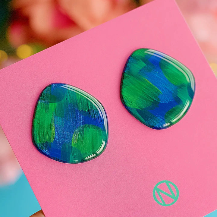 The Peacock Vibes Stud Earrings by Naoi are artistically designed with irregular shapes and vibrant green and blue brushstrokes. Hand-painted and featuring stainless steel posts, these unique jewelry pieces are displayed on a pink card with a minimalist "N" logo within a circle in the bottom right corner. The background is softly blurred.