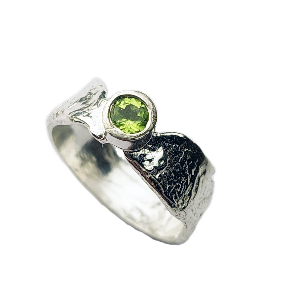 A silver ring featuring a textured, uneven surface with a prominent round green gemstone set in the center. Dubbed the "Faerie Tale Ring Thin Band" by Caroline Stokesberry-lee, the band has a rough, organic appearance, giving it a unique, artisanal look.