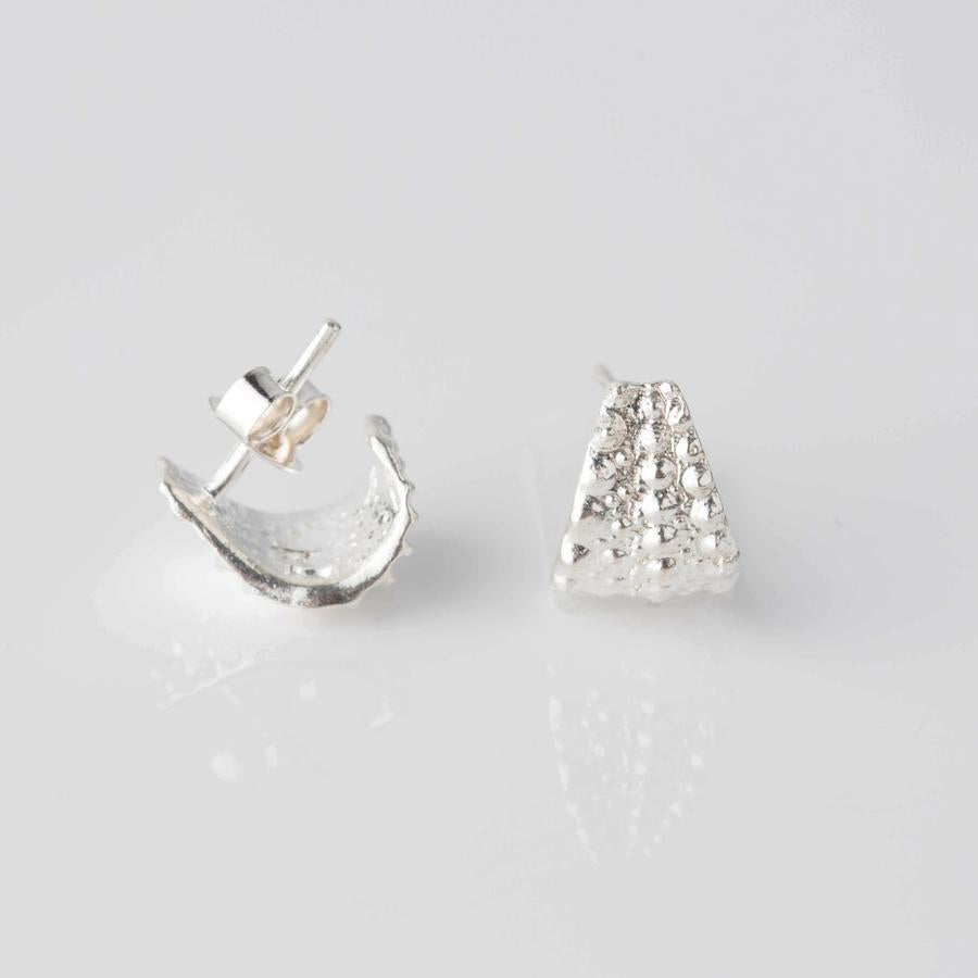 A pair of elegant everyday studs, with one lying flat showing a textured triangular front and the other tilted on its side revealing a textured curved edge. These timeless Sea Urchin Wrap Stud Earrings by Jennifer Kinnear are both placed against a plain white background.