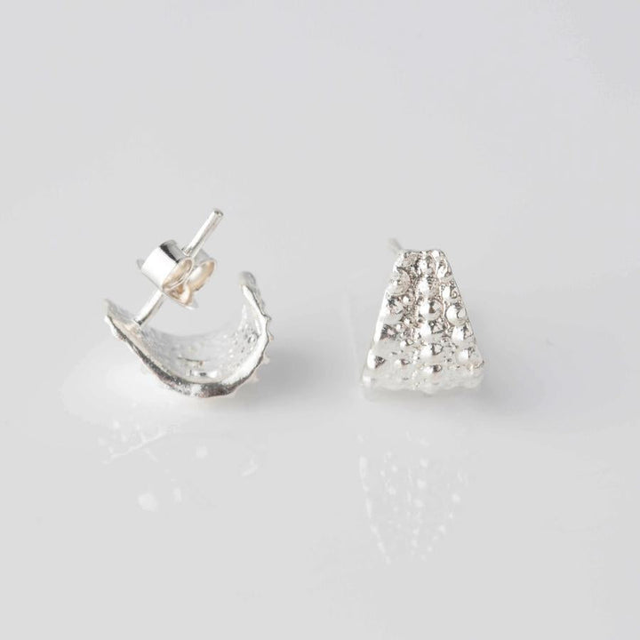 A pair of elegant everyday studs, with one lying flat showing a textured triangular front and the other tilted on its side revealing a textured curved edge. These timeless Sea Urchin Wrap Stud Earrings by Jennifer Kinnear are both placed against a plain white background.