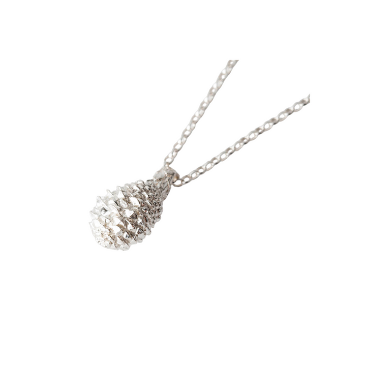 A close-up image of a silver necklace featuring a pendant shaped like a small, intricate baby pine cone. The delicate chain with interconnecting small links highlights the shiny, textured **Pine Cone Long Necklace** by **Jennifer Kinnear** hanging at the center. The background is white.