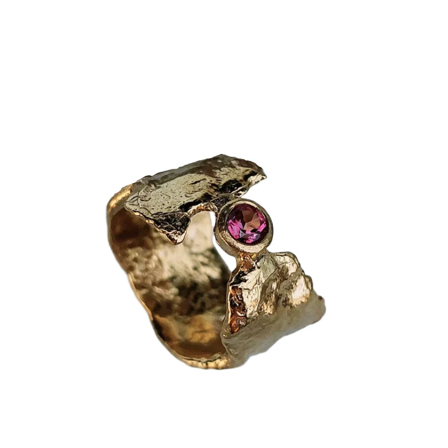 The Pink Topaz & Gold Faerie Tale Ring, a yellow gold plated textured piece with a jagged design from Caroline Stokesberry-lee, features a captivating pink topaz gemstone. Displayed against a blurred background of text and twisted metal wire, its rough, organic shapes meld effortlessly with the elegance of the gemstone.