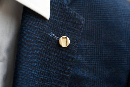 Close-up of a person wearing a blue checkered suit jacket with a Millett Wade Pint Lapel Pin on the lapel. The white shirt collar is visible above the lapel. The suit has a textured pattern and the pin is small and round.