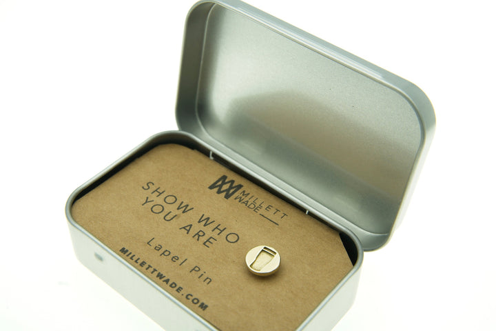 A small, hand-finished brass lapel pin is displayed inside an open silver tin box. The pin is attached to a brown card that reads, "Millett Wade, Show Who You Are, Pint Lapel Pin." The website "millettmade.com" is also printed on the card.