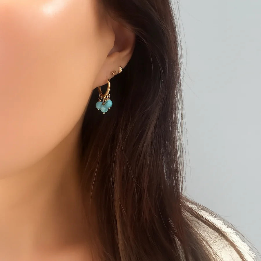 Close-up of a woman's ear with long brown hair, wearing Aria V’s Polly Earrings—small gold-plated hoops adorned with three light blue beads—and a triangular stud. Elegant handmade Irish-style jewelry shown against a plain light gray background.