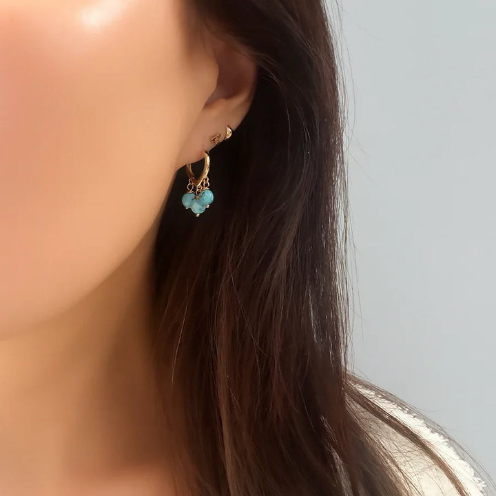 Close-up of a woman's ear with long brown hair, wearing Aria V’s Polly Earrings—small gold-plated hoops adorned with three light blue beads—and a triangular stud. Elegant handmade Irish-style jewelry shown against a plain light gray background.
