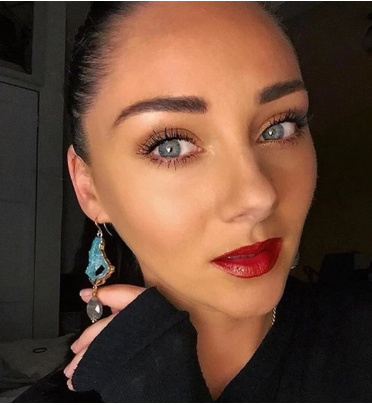 Close-up of a person with light skin, blue eyes, dark hair pulled back, wearing red lipstick, and intricate handcrafted Cameo Earrings by Aria V adorned with turquoise teardrops. They are looking directly at the camera with a slight tilt of the head, and wearing a black top. The background is slightly blurred.