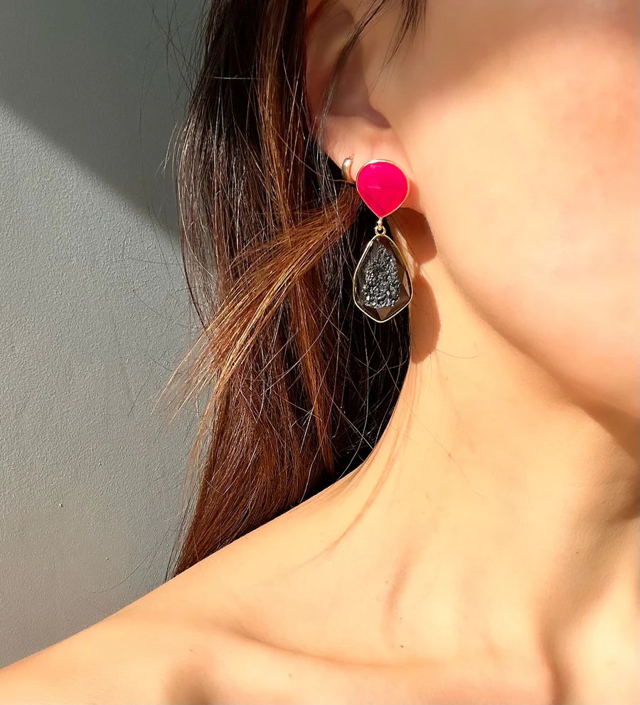 Close-up of a person's ear adorned with Poppy Earrings. The earring features a bright pink triangular stud connected to a dangling teardrop-shaped design with a textured chalcedony stone in the center, set against a gold-toned frame. These exquisite earrings are by Aria V.