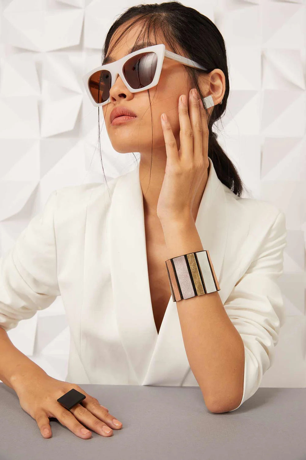A woman with long dark hair, wearing white-framed sunglasses and a white blazer, sits with her hand resting on her cheek. She wears the Rainbow Round Bracelet in metallic colors from Iskin Sisters and a matching ring made of lightweight jewelry. The background features a geometric white pattern.